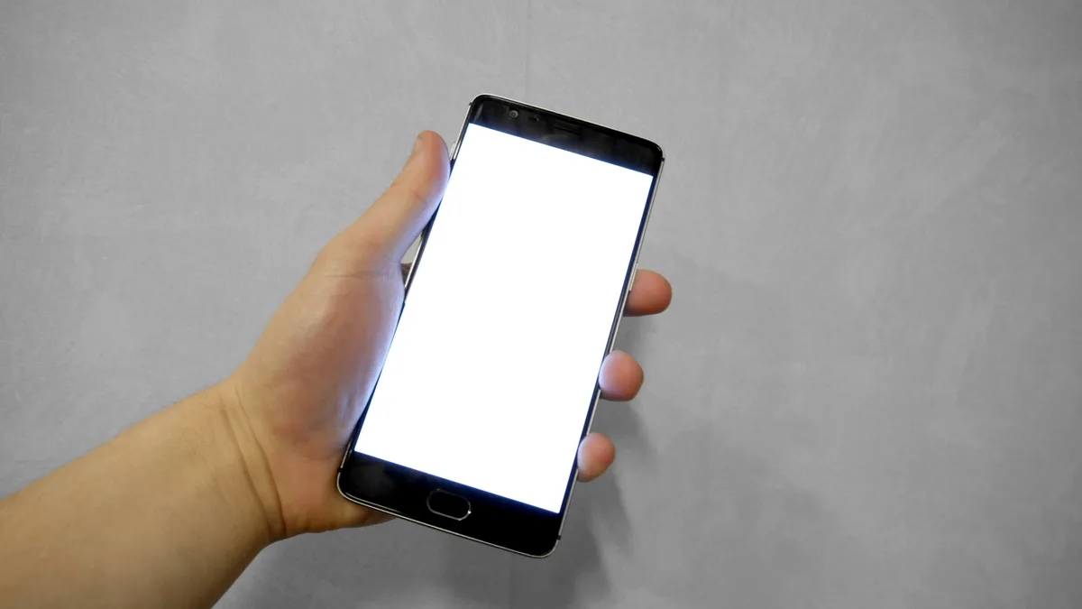 A hand holding a smartphone with a blank white screen in front of a grey wall